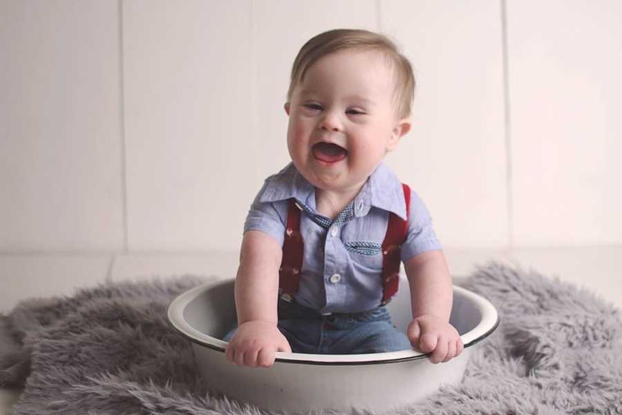 Baby with down syndrome smiles as he wears overalls sitting in bucket