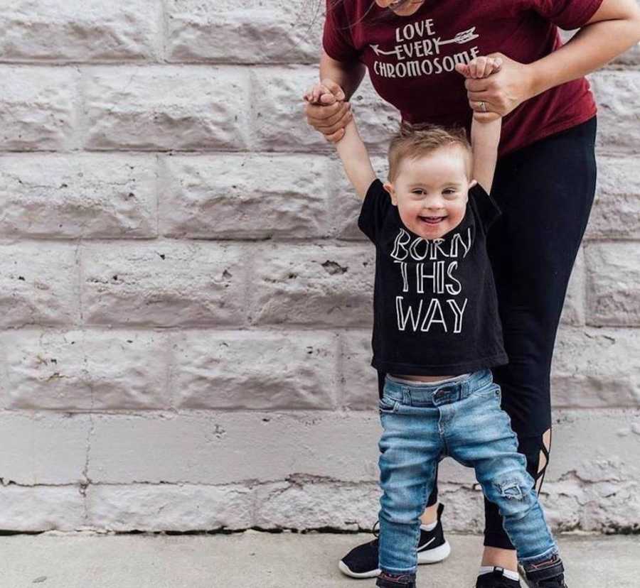 Little boy with down syndrome stands wearing shirt that says, "born this way" as mother holds his hands