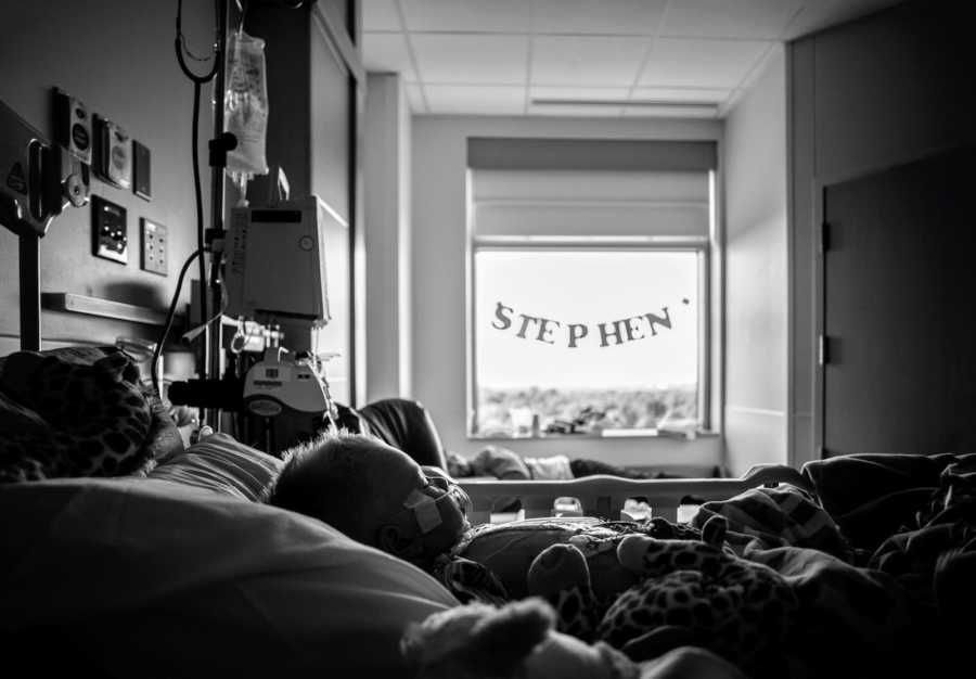 Little boy with congenital heart defects lays in hospital bed as window in room has sign that says, "Stephen"
