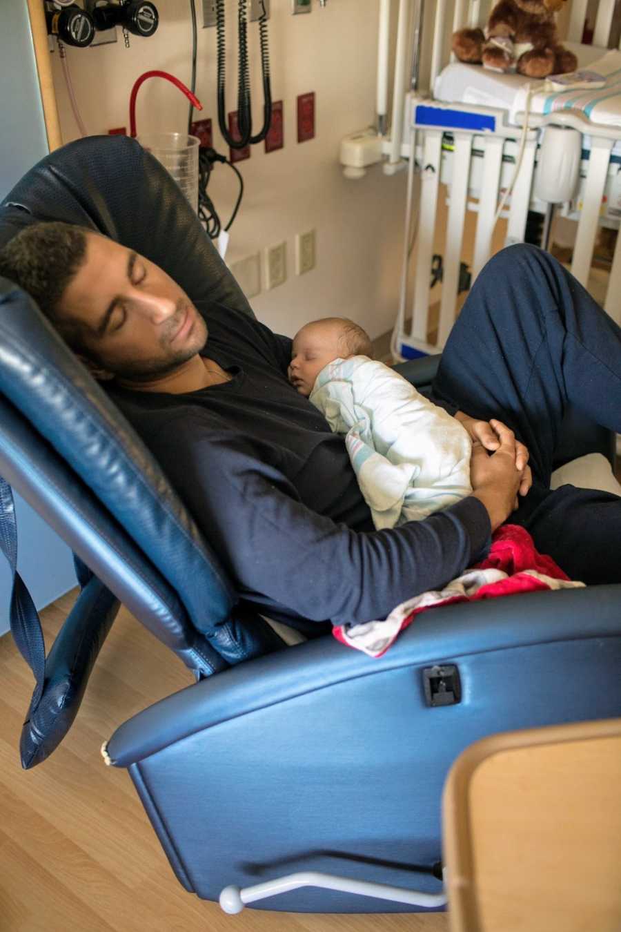 Father sits in hospital chair asleep with baby as he waits for other son to wake up from heart surgery