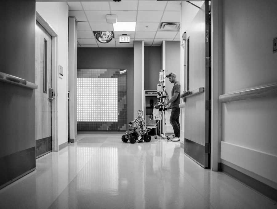 Little boy rides toy four wheeler in hospital hallway while father pushed monitor behind him