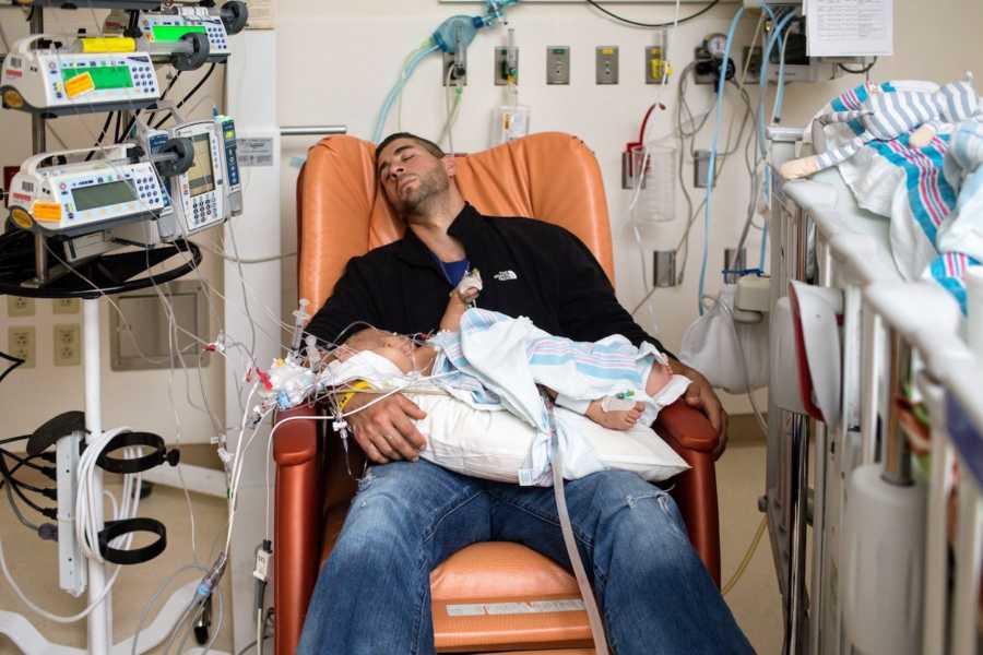 Father sits asleep in hospital chair while baby son who just had open heart surgery lays in his lap