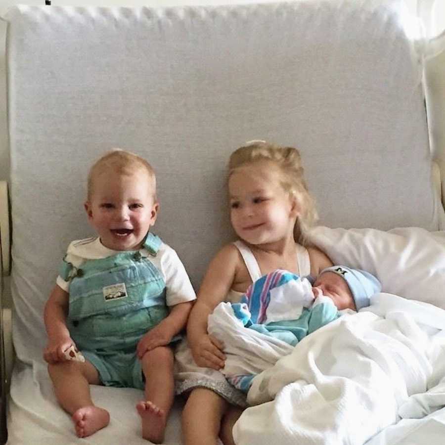 Brother and sister sit smiling as sister has baby sibling in her lap