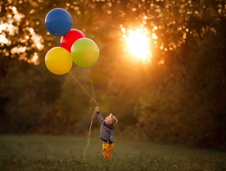 Little boy with congenital heart defects stands outside holding string of balloons