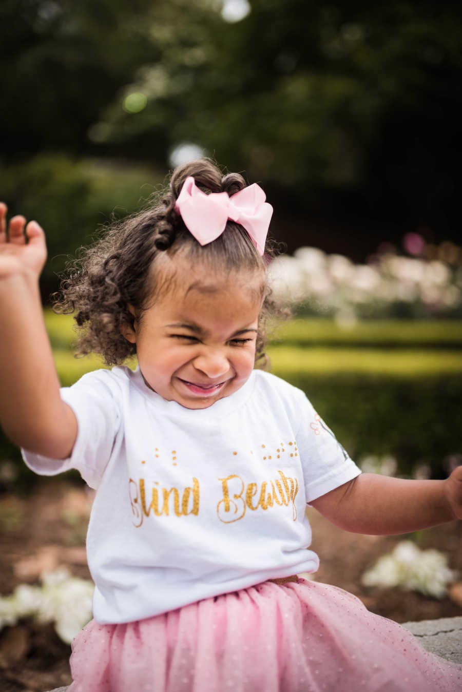 Little girl with prosthetic eyes sits smiling outside with shirt that says, "blind beauty"