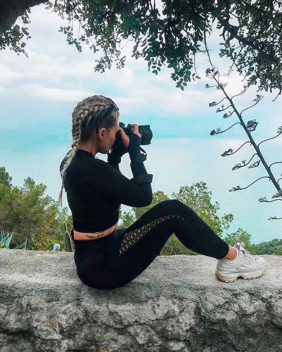 Woman with BPD and CPTSD sits on ledge holding camera as she looks out at plants and body of water