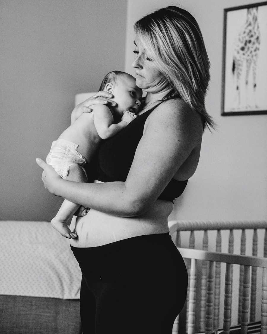 Mother with eating disorder who has gained weight stands in sports bra and leggings holding baby