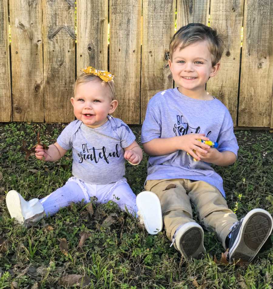 Young boy sits on ground outside in yard beside his little sister