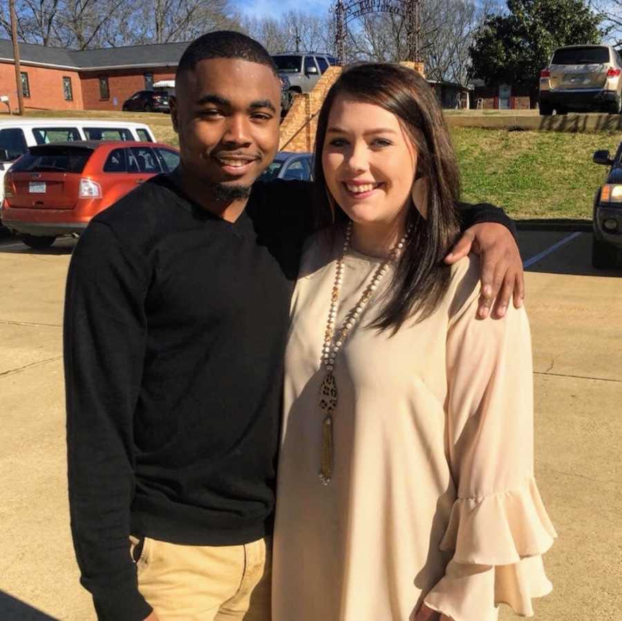 Boyfriend and girlfriend of different races stand smiling in parking lot with arms around each other