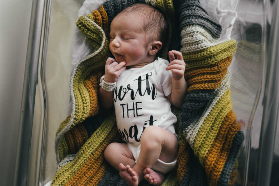 Newborn baby lays on knit blanket wearing onesie that says, "worth the wait"