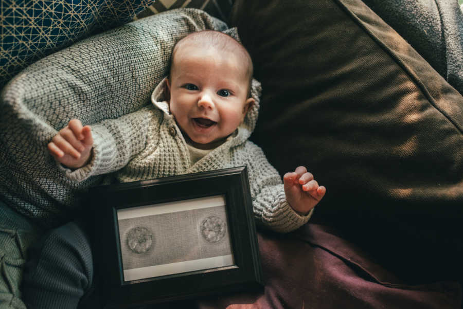 Baby smiles as he lays on couch with picture frame of implanted embryos on his chest