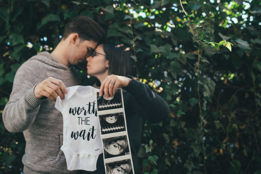Husband and wife stand outside with their heads together as they hold onesie and sonogram pictures