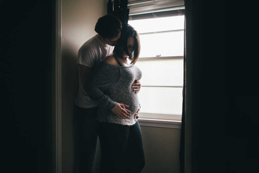 Husband stands behind pregnant wife hugging her in home