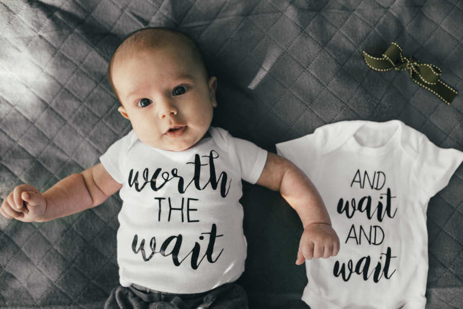 Newborn lays on back wearing onesie that says, "worth the wait" beside onesie for his younger sibling