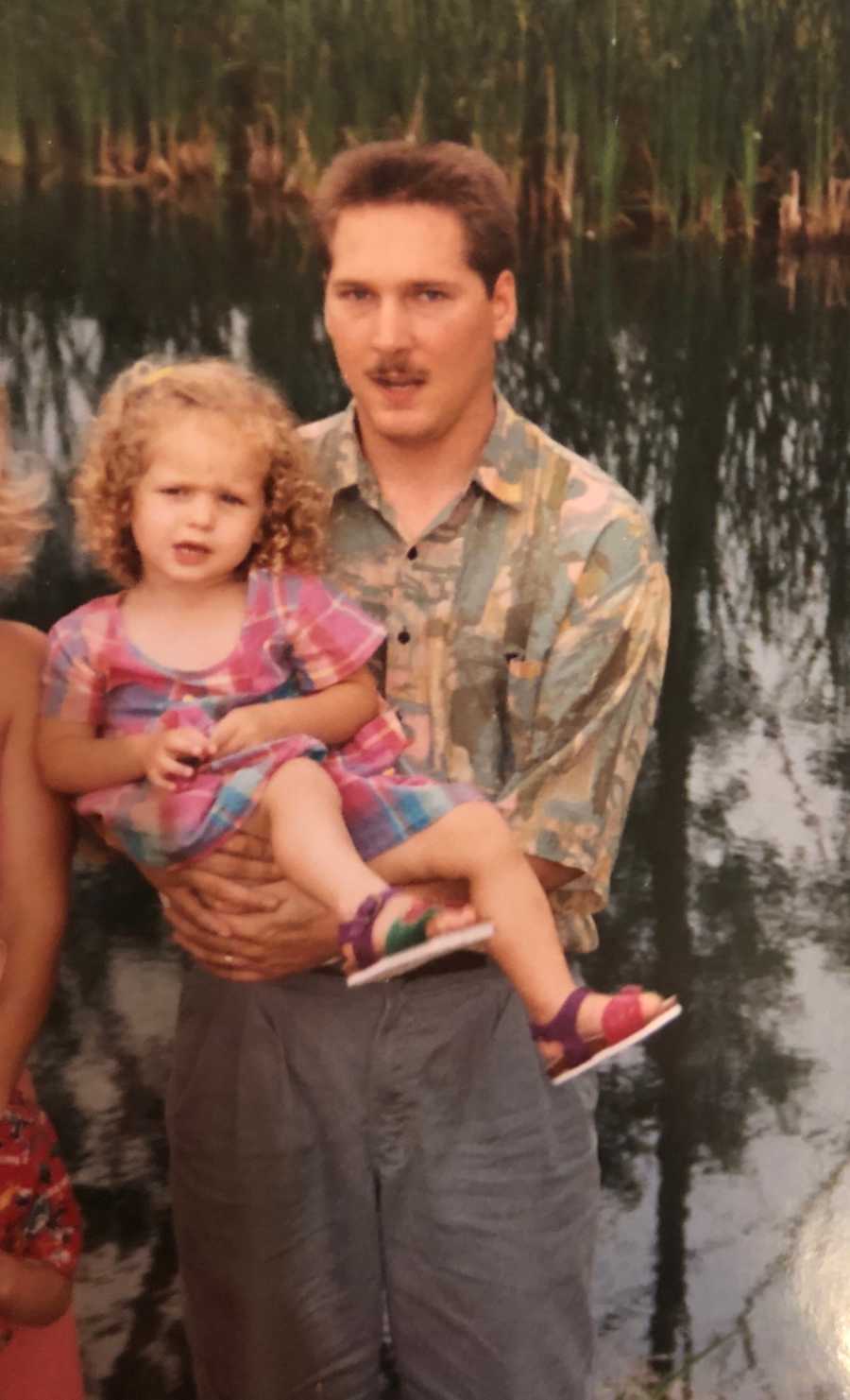 Man who has since passed stands near body of water holding his young daughter