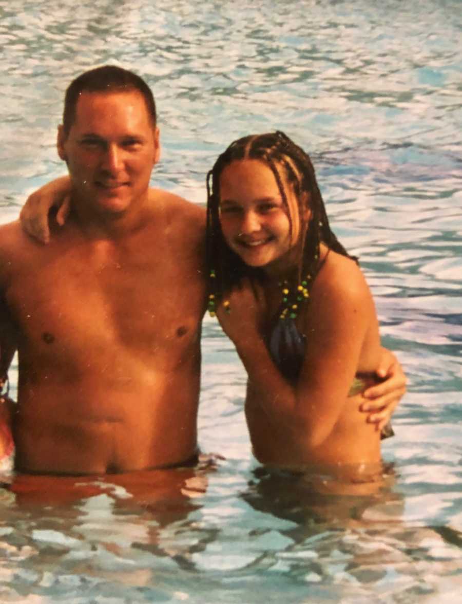 Man smiles in body of water with his arm around his teen daughter