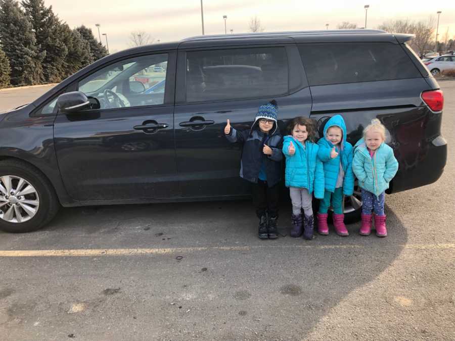 Four little kids stand in parking lot in front of their van with thumbs up
