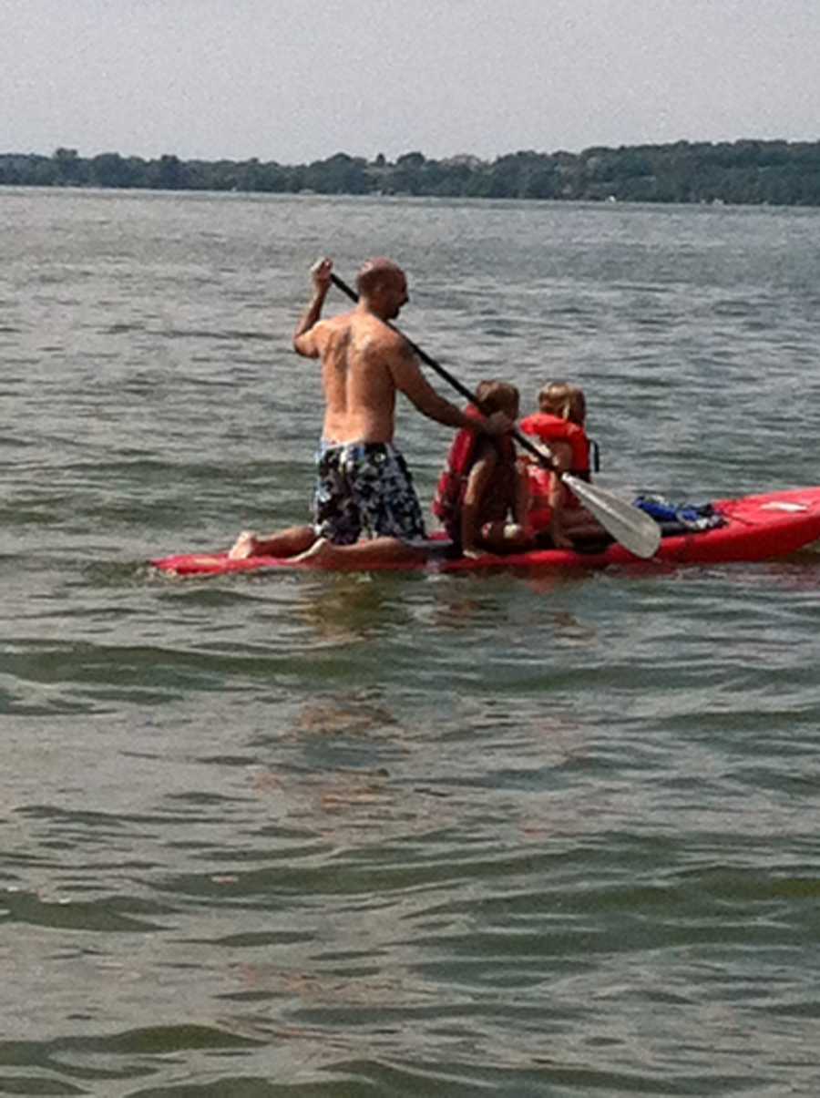Father kneels on paddle board paddling as his two kids sit in front of him