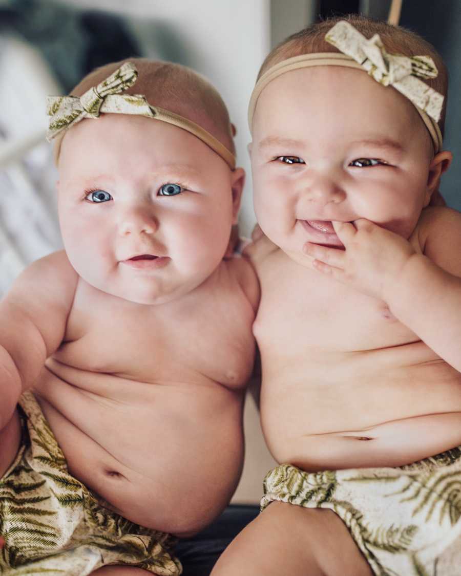 Twin baby girls sits smiling in matching bloomers and headband