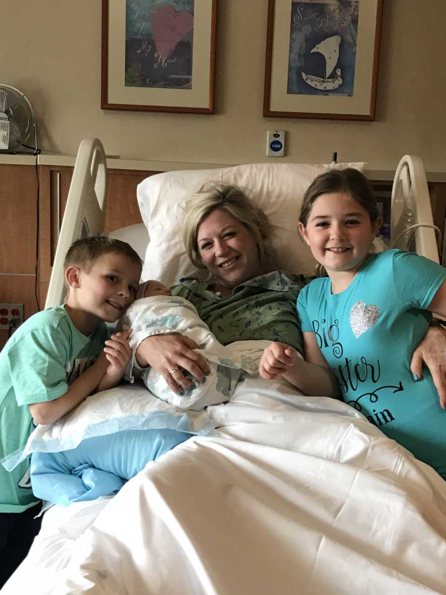 Widow lays in hospital bed holding newborn while her other two kids stand on either side of bed