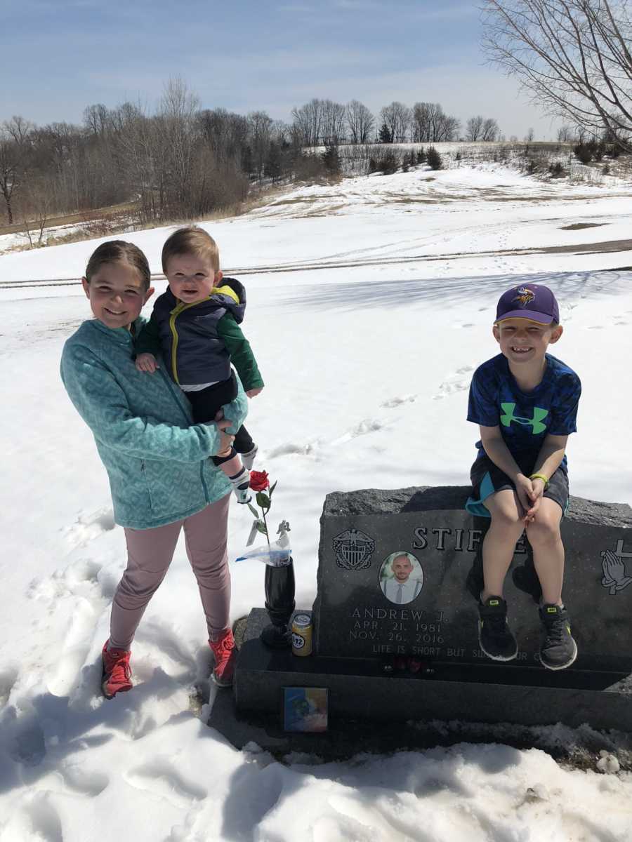 Little girl holds youngest sibling beside father's headstone while their brother sits on it