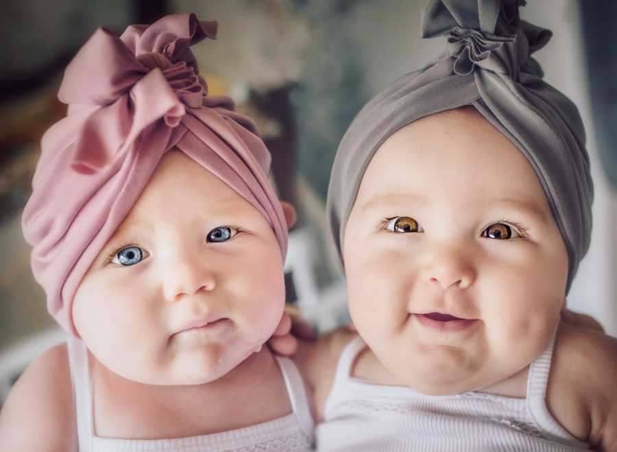 Twin baby girls sit beside each other wearing pink and grey head wrap