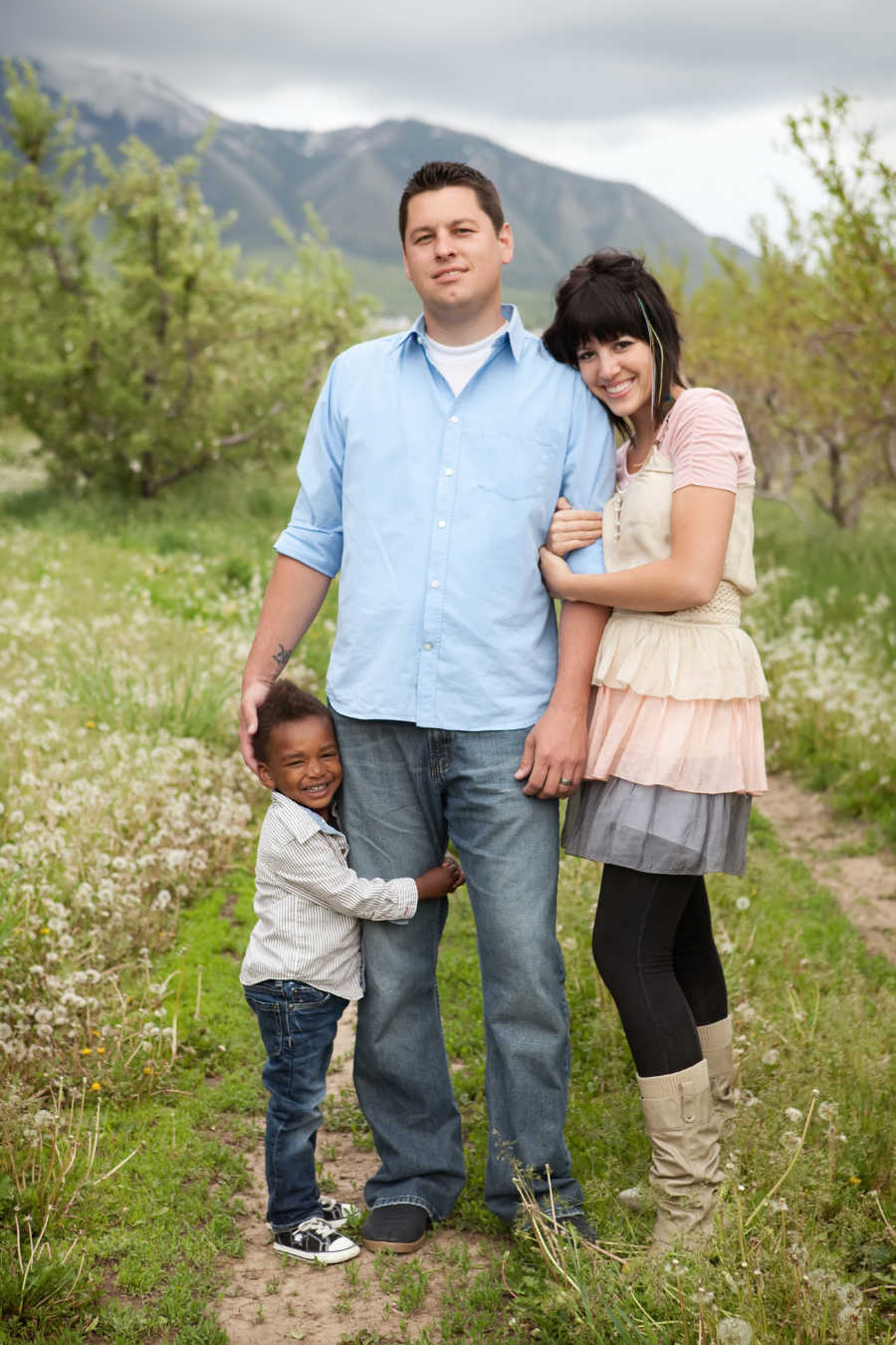 Husband and wife stand outside while their adopted son stands holding on to father's leg
