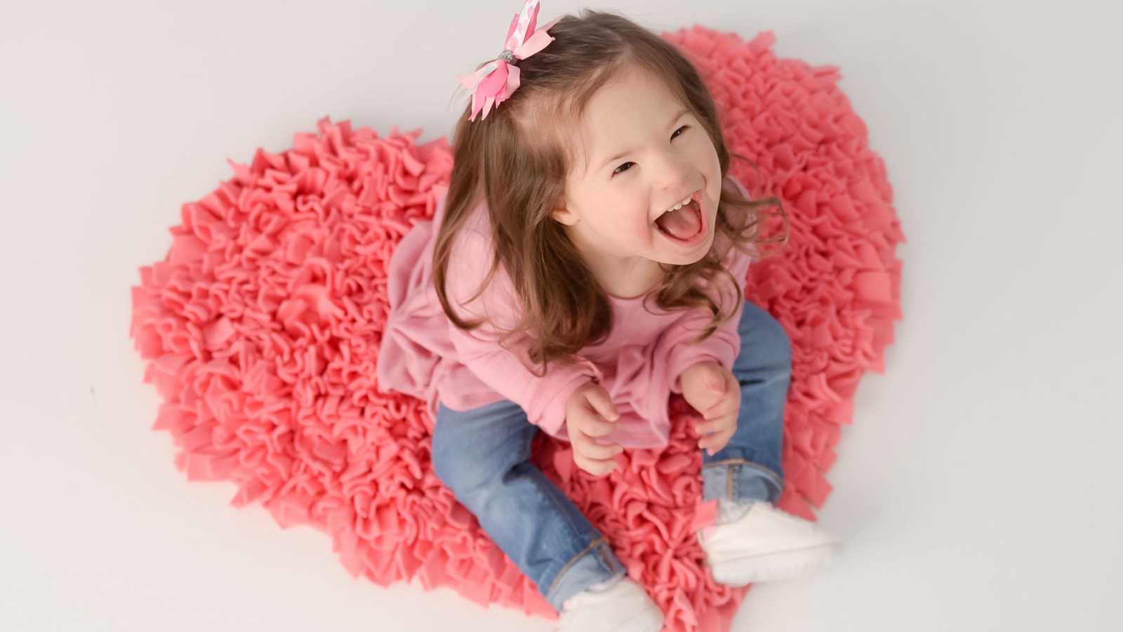 Little girl with down syndrome sits on heart shaped rug as she looks up smiling