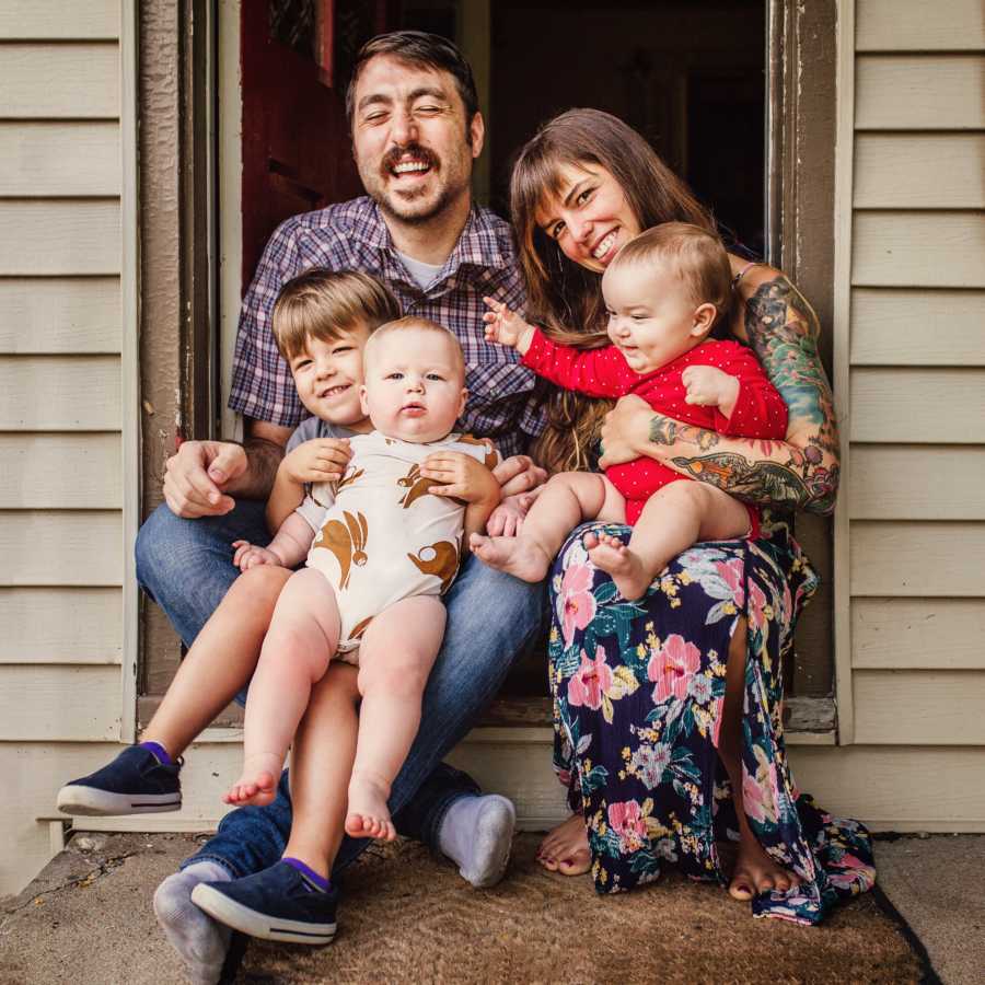 Husband and wife sit on step of their front door with their three kids in their lap