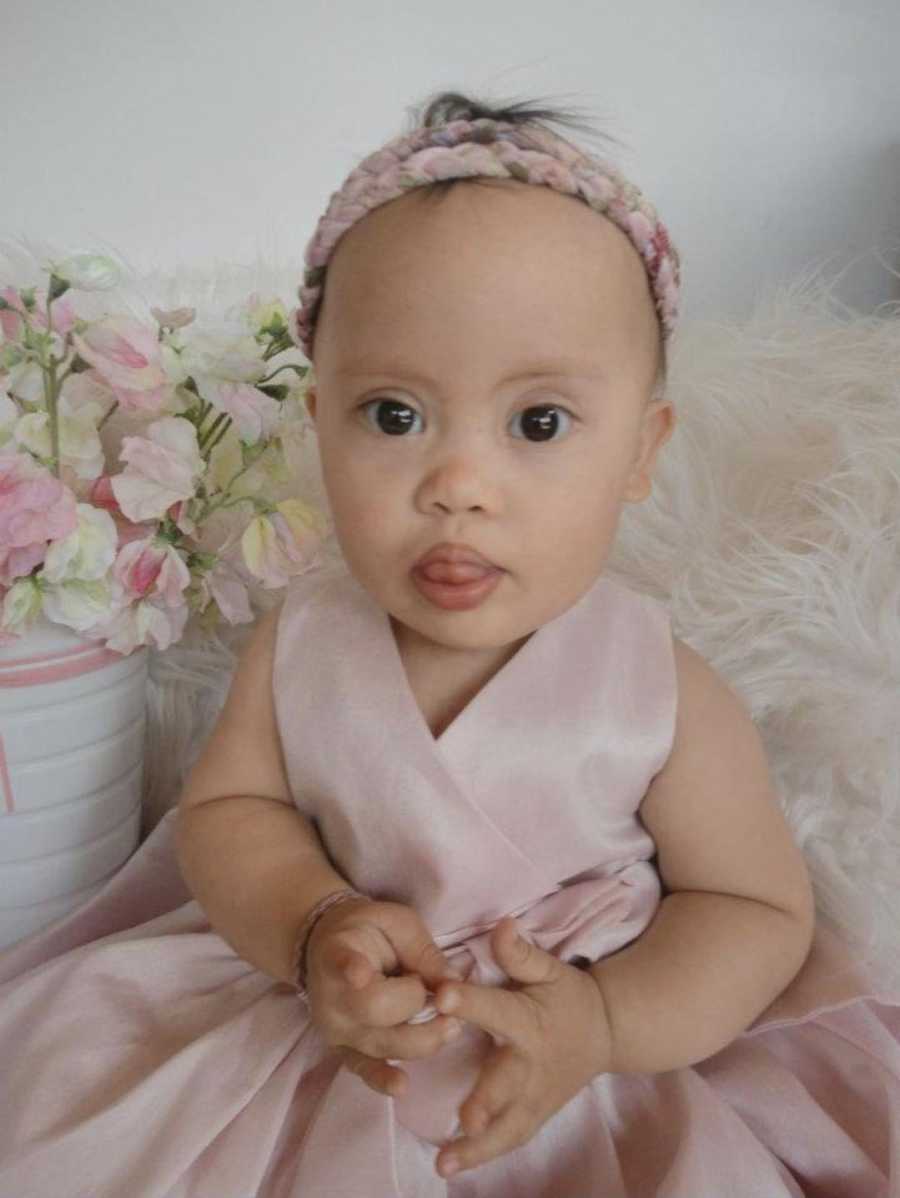 Baby with light pink dress on sits with her tongue out