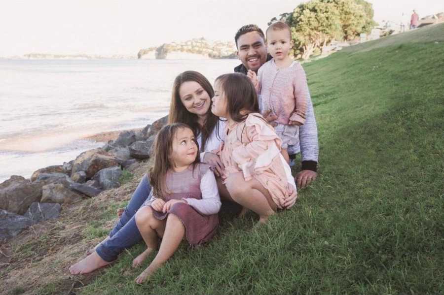 Husband and wife sit on grass near body of water with their three young daughters