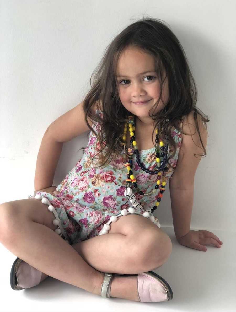 Little girl who has tumor removed from brain sits smiling with floral dress on