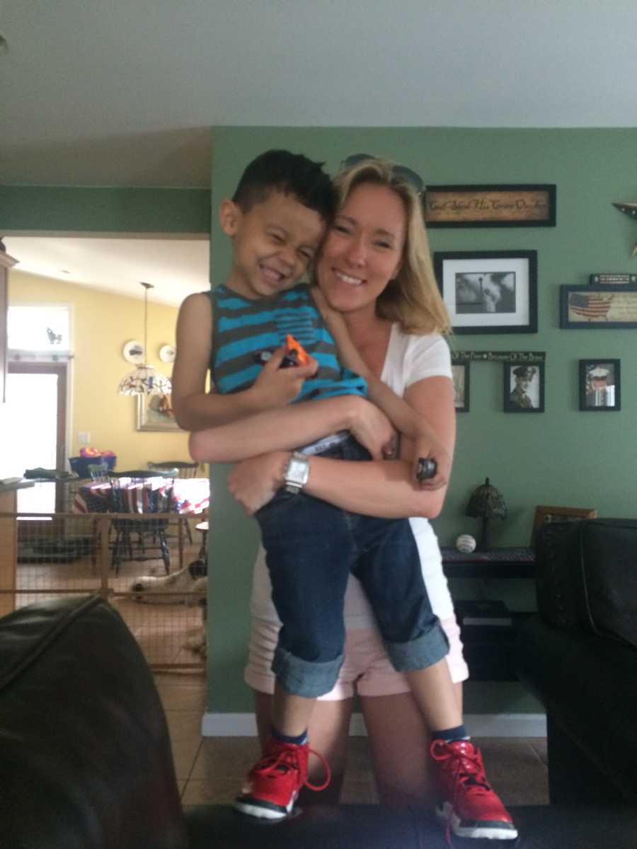 Woman stands smiling in home while she hugs boyfriend's son who stands on couch