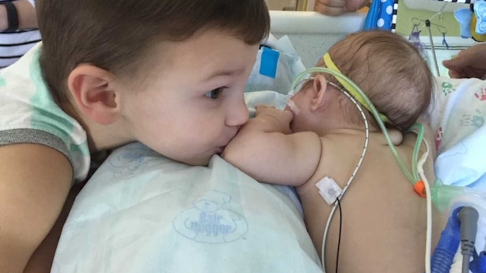 Little boy kisses arm of baby sister who lays in NICU after open heart surgery