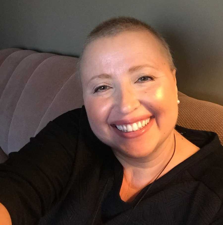 Woman with breast cancer and shaved head smiles in selfie as she sits on couch in home