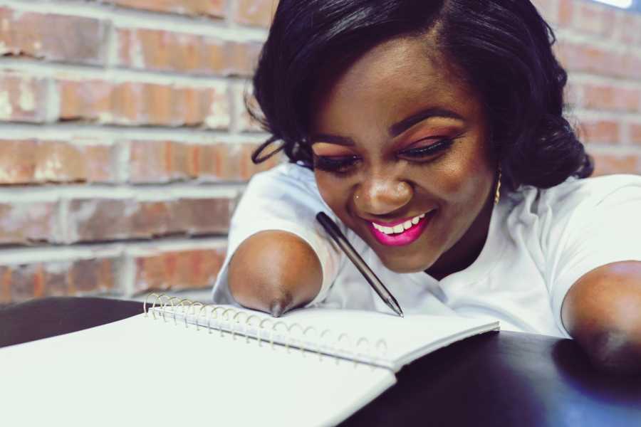 Woman without bottom half of arms smiles as she sits at table with notebook and pen