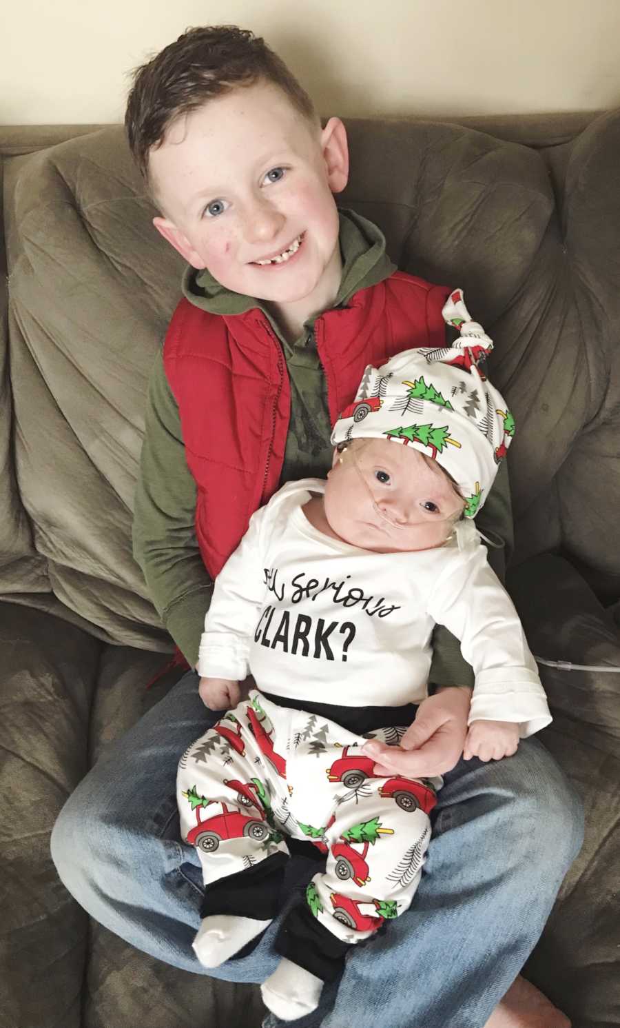 Little boy smiles as he sits on couch holding baby brother on oxygen in his lap