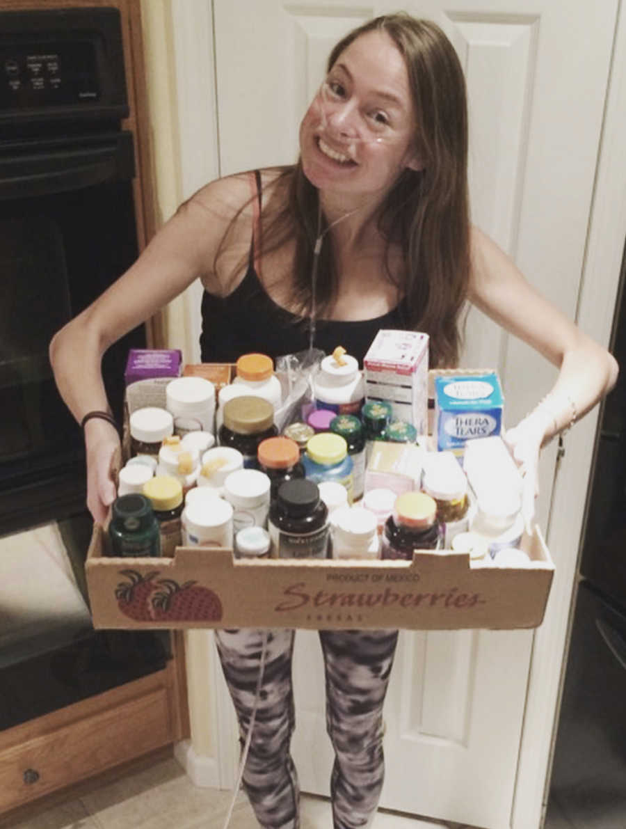 Woman with Cystic Fibrosis and on oxygen stands in home smiling as she holds box of medications