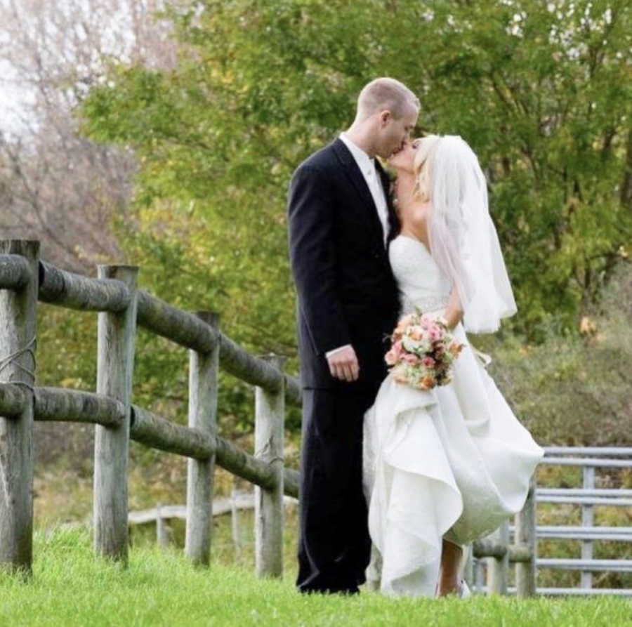 Bride and groom stand outside by wooden fence kissing each other