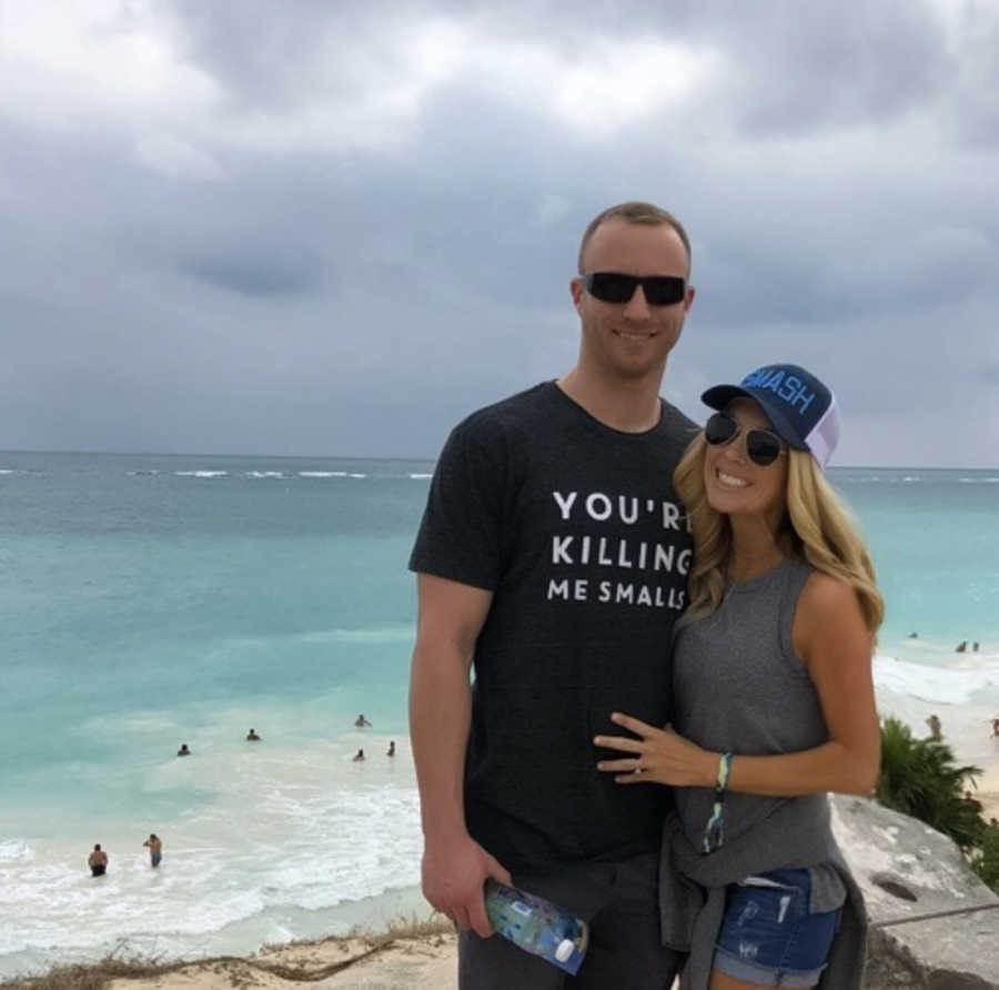 Husband and wife stand outside smiling with ocean in background