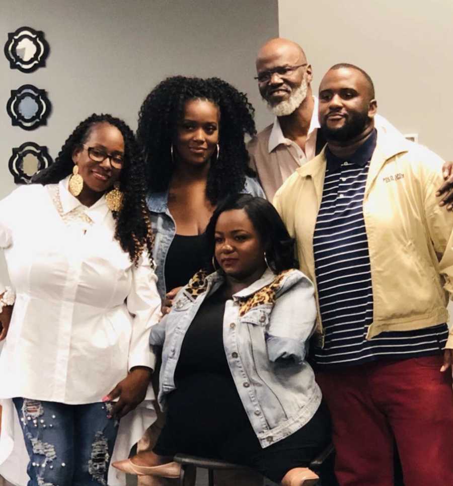 Woman without bottom half of arms smiles as she sits on stool with her family members behind her