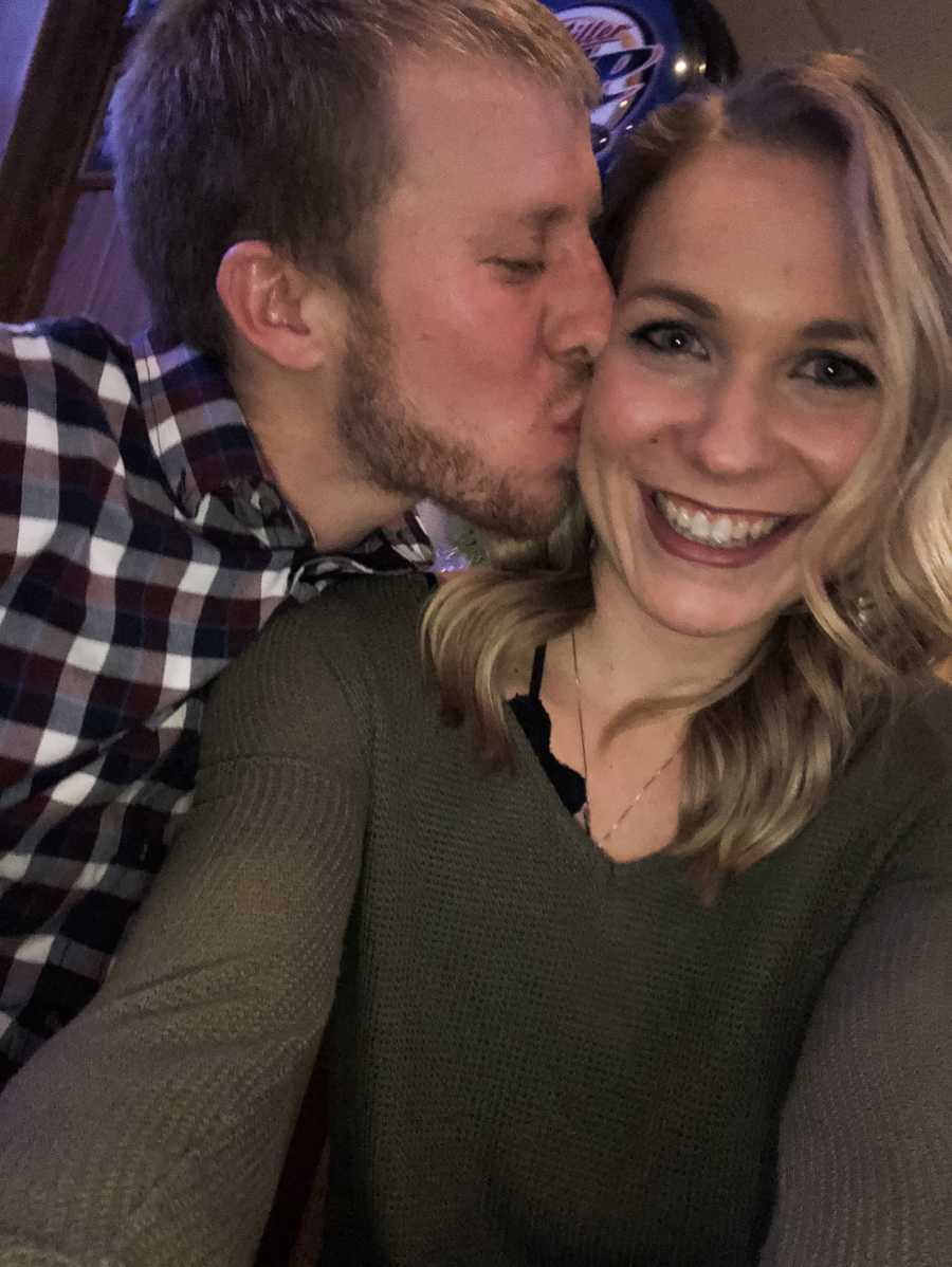 Woman smiles in selfie as her husband kisses her on the cheek