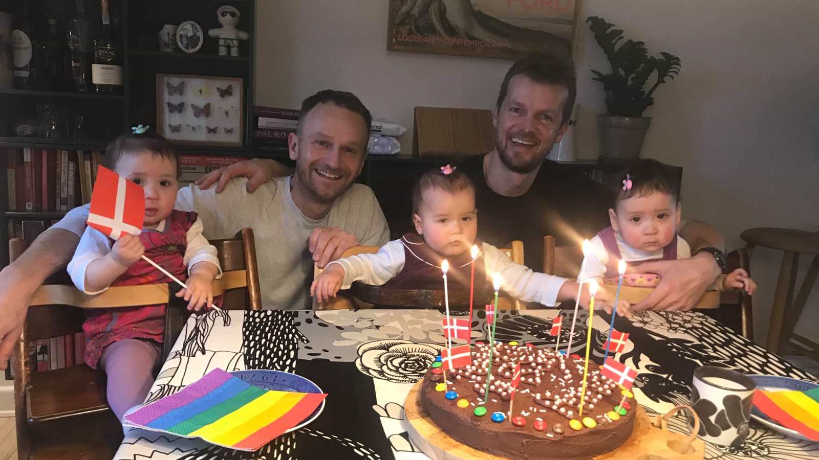Husbands smiles as they crouch behind their triplets in highchairs who sit at table with birthday cake