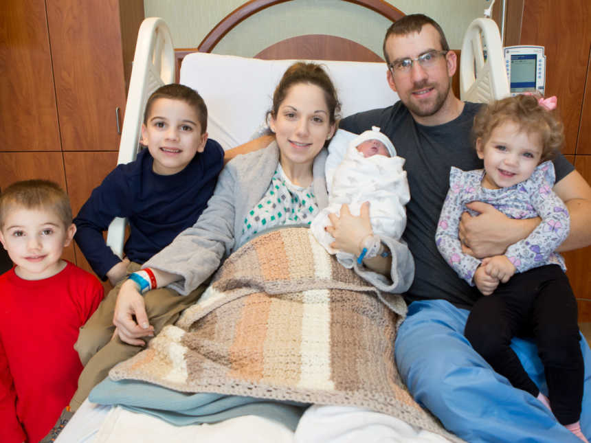Husband and wife sit in bed with their newborn and their three other kids