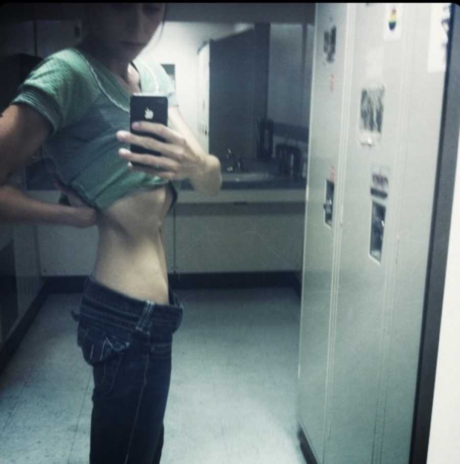 Woman with eating disorder stands in locker room taking mirror selfie with shirt pulled up