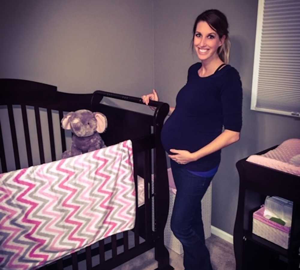 Pregnant woman stands smiling beside crib in home