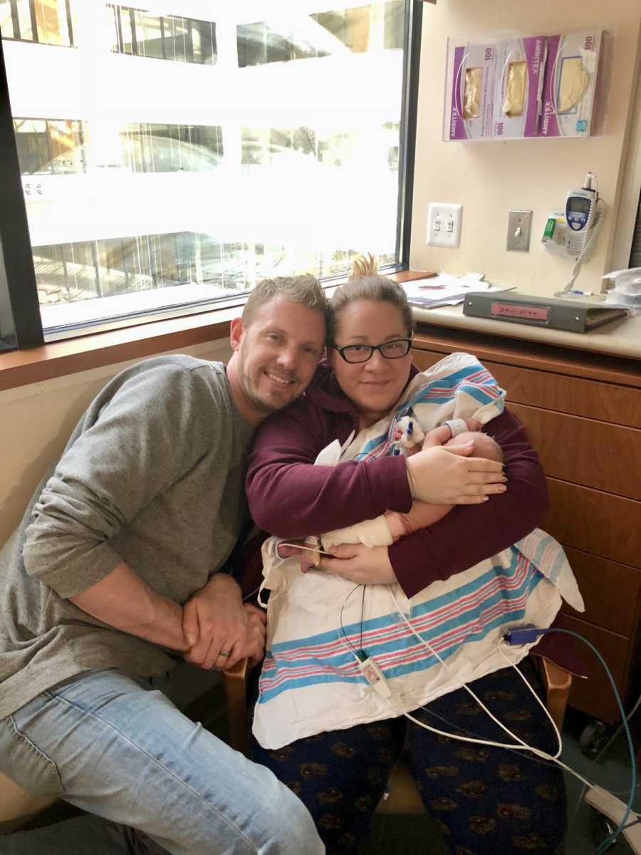 Mother sits in NICU holding newborn in her arms as her husband sits beside her