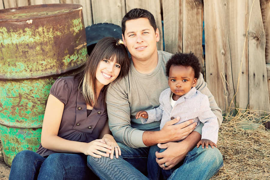 Husband and wife sit on ground outside as husband has their adopted child in his lap