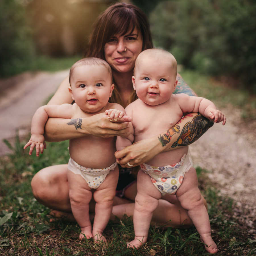 Mother smiles as she sits on ground outside holding twin babies that stand in her arms