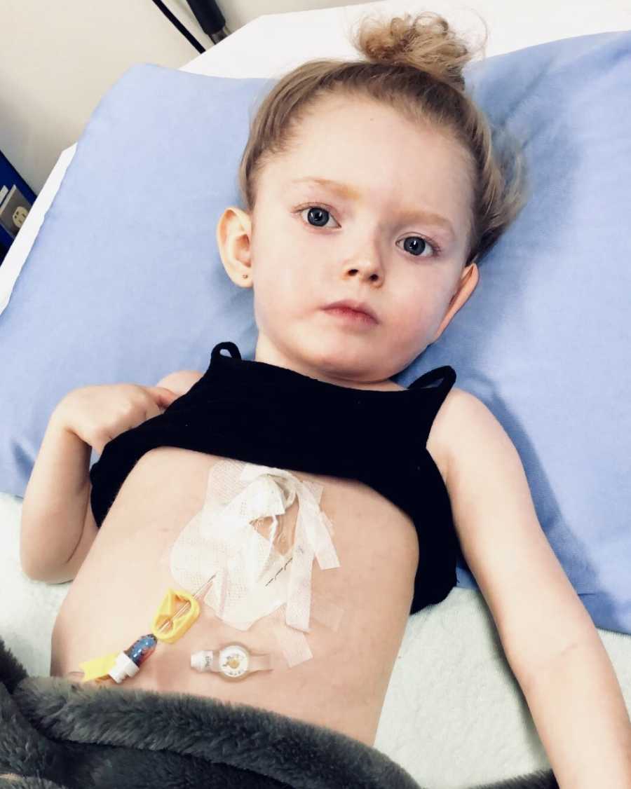 Little girl with Rett Syndrome lays on back in hospital with shirt lifted to show feeding tube